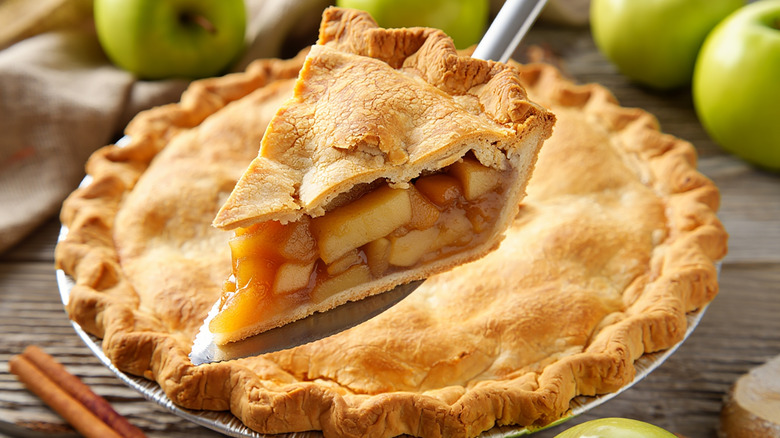 Close-up of a slice of apple pie being held on a pie spatula above a whole pie with green apples and cinnamon sticks blurred around it