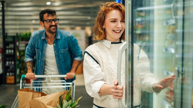 A couple pushes a cart and reaches into a freezer to grab something from the grocery store