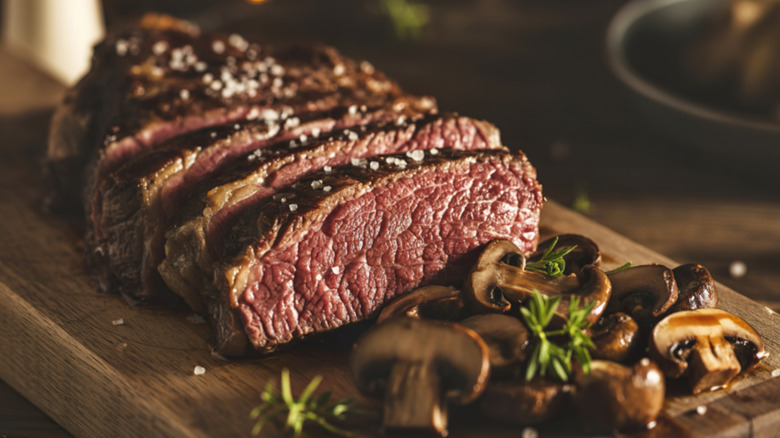 A thick medium-rare ribeye steak, sliced with mushrooms on a cutting board