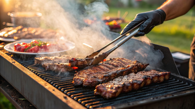 A gloved hand with tongs grilling steaks
