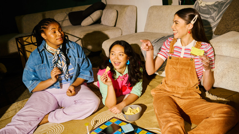 A group of young women in 1980s attire eating snacks while playing a board game